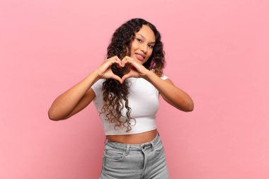 Genç İspanyol kadın gülümsüyor ve mutlu, sevimli, romantik ve aşık, iki eliyle kalbi şekillendiriyor.