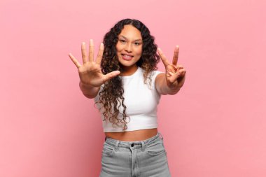 Genç İspanyol kadın gülümsüyor ve dost canlısı görünüyor, 7 ya da 7 numarayı gösteriyor, eli önde, geri sayıyor.