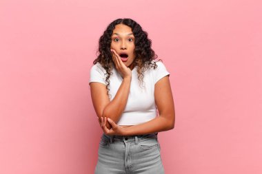 Genç İspanyol kadın şok ve inançsızlıkla ağzı açık, elleri yanak ve kolları çapraz, sersemlemiş ve şaşırmış hissediyor.