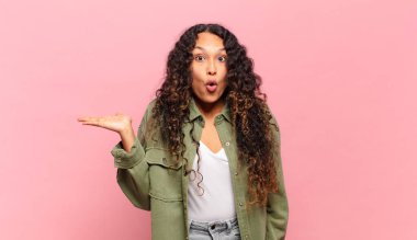 Genç İspanyol kadın şaşırmış ve şoka uğramış görünüyor, çenesi düşmüş, eli açık bir objeyi tutuyor.