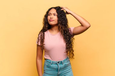 Genç İspanyol kadın şaşkın ve şaşkın hissediyor, kafasını kaşıyor ve yan tarafa bakıyor.