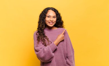 Genç İspanyol kadın neşeyle gülümsüyor, mutlu hissediyor ve kenarı ve yukarıyı işaret ediyor, fotokopi alanında nesneler gösteriyor.