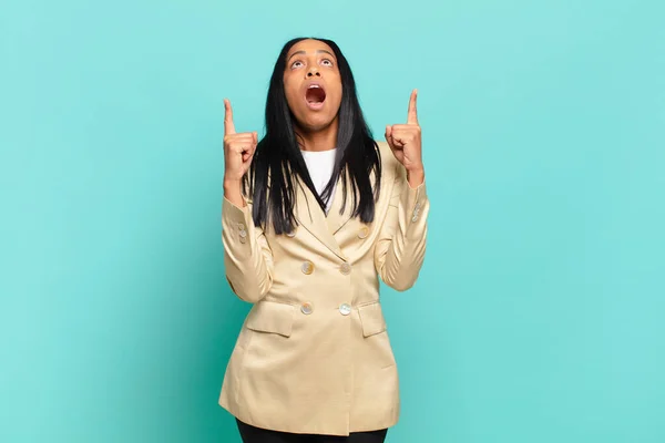 young black woman looking shocked, amazed and open mouthed, pointing upwards with both hands to copy space. business concept