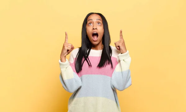 young black woman looking shocked, amazed and open mouthed, pointing upwards with both hands to copy space