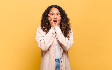 Genç İspanyol kadın şok olmuş ve korkmuş hissediyor, ağzı açık ve elleri yanaklarında korkmuş görünüyor.