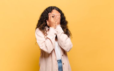 Latin kökenli genç bir kadın korkmuş ya da utanmış hissediyor. Gözleri yarı kapalı ya da elleri kapalı gözetliyor.