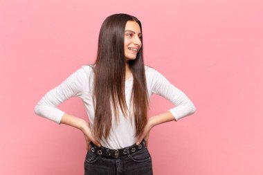 Genç, güzel bir kadın mutlu, neşeli ve kendinden emin görünüyor. Gururla gülümsüyor ve iki elini de kalçasında istiyor.