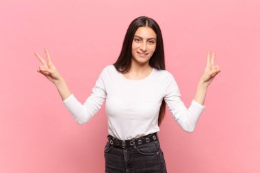 Genç ve güzel bir kadın gülümsüyor ve mutlu görünüyor, arkadaş canlısı ve memnun, işaret eden zafer ya da barış iki elle