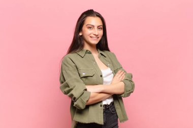 Genç ve güzel bir kadın kollarını kavuşturup mutlu bir şekilde gülüyor, rahat, pozitif ve tatmin edici bir pozla.