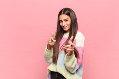 Genç, güzel bir kadın, olumlu, başarılı, mutlu bir tavırla kamerayı işaret ederek, elleriyle silah işareti yaparak gülümsüyor.