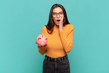 Genç ve güzel bir kadın şok olmuş ve korkmuş hissediyor, ağzı açık ve elleri yanaklarında korkmuş görünüyor. Domuz kumbarası konsepti
