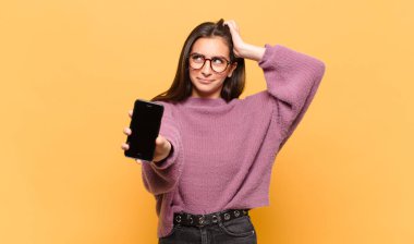 Genç, güzel bir kadın şaşkın ve kafası karışmış hissediyor. Kafasını kaşıyor ve yan tarafa bakıyor. telefon ekranı kavramı