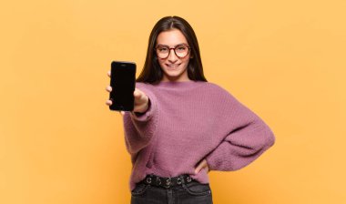 Genç ve güzel bir kadın mutlu bir şekilde gülümsüyor, eli kalçasında ve kendinden emin, olumlu, gururlu ve arkadaş canlısı bir tavırla. telefon ekranı kavramı