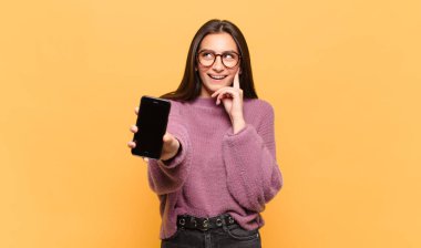 Genç ve güzel bir kadın mutlu bir şekilde gülümsüyor ve hayal kuruyor ya da şüphe ediyor, yan tarafa bakıyor. telefon ekranı kavramı