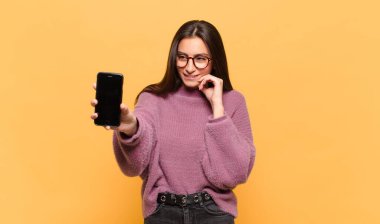 Genç, güzel bir kadın, mutlu, kendinden emin bir ifadeyle gülümsüyor, elleri çenesinde, merak ediyor ve yan tarafa bakıyor. telefon ekranı kavramı