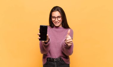 Genç, güzel bir kadın gururlu, tasasız, kendinden emin ve mutlu, başparmağıyla olumlu gülümsüyor. telefon ekranı kavramı