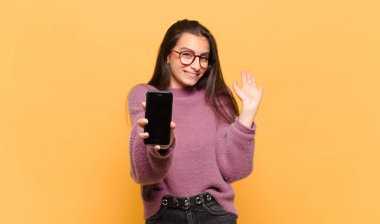 Genç ve güzel bir kadın mutlu ve neşeli bir şekilde gülümsüyor, el sallıyor, sizi selamlıyor ve selamlıyor, ya da veda ediyor. telefon ekranı kavramı
