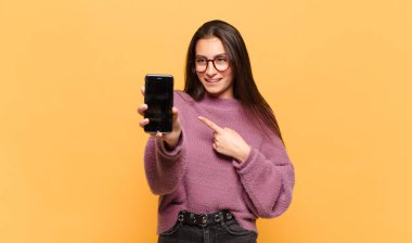 Genç, güzel bir kadın neşeyle gülümsüyor, mutlu hissediyor ve yan ve yukarı doğru işaret ediyor, fotokopi uzayında nesneler gösteriyor. telefon ekranı kavramı