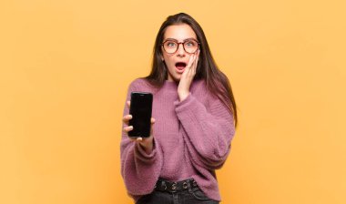 Genç ve güzel bir kadın şok olmuş ve korkmuş hissediyor, ağzı açık ve elleri yanaklarında korkmuş görünüyor. telefon ekranı kavramı