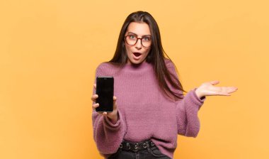 Genç ve güzel bir kadın şaşırmış ve şok olmuş görünüyor. Çenesi düşmüş ve eli açık bir objeyi tutuyor. telefon ekranı kavramı