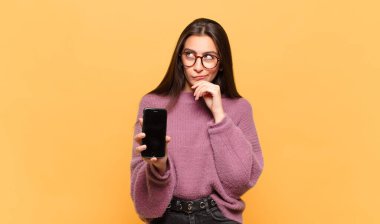 Genç, güzel bir kadın düşünüyor, kuşkulu ve şaşkın hissediyor, farklı seçeneklerle, hangi kararı vereceğini merak ediyor. telefon ekranı kavramı