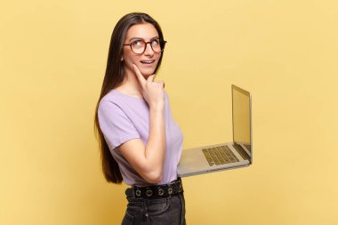 Genç ve güzel bir kadın mutlu bir şekilde gülümsüyor ve hayal kuruyor ya da şüphe ediyor, yan tarafa bakıyor. dizüstü bilgisayar