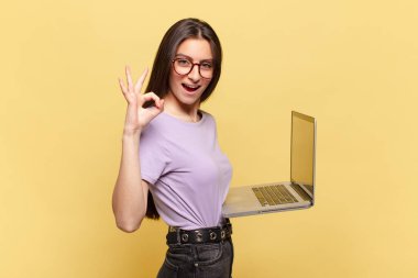 Genç, güzel bir kadın kendini mutlu, rahat ve tatmin olmuş hissediyor. İyi bir jestle onaylıyor, gülümsüyor. dizüstü bilgisayar