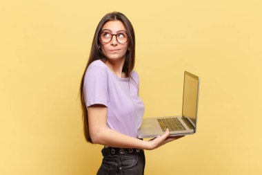 Genç, güzel bir kadın omuz silkiyor, kafası karışık ve kararsız, kolları bağlı ve şaşkın görünüyor. dizüstü bilgisayar