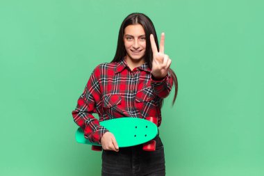 Genç ve güzel bir kadın gülümsüyor ve mutlu görünüyor, kaygısız ve pozitif, tek elle zafer ya da barış işareti yapıyor. kaykay konsepti