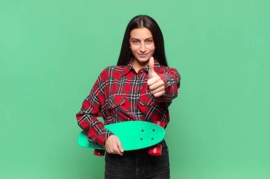 Genç, güzel bir kadın gururlu, tasasız, kendinden emin ve mutlu, başparmağıyla olumlu gülümsüyor. kaykay konsepti