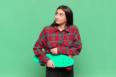 Genç, güzel bir kadın omuz silkiyor, kafası karışık ve kararsız, kolları bağlı ve şaşkın görünüyor. kaykay konsepti