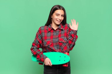 Genç ve güzel bir kadın mutlu ve neşeli bir şekilde gülümsüyor, el sallıyor, sizi selamlıyor ve selamlıyor, ya da veda ediyor. kaykay konsepti