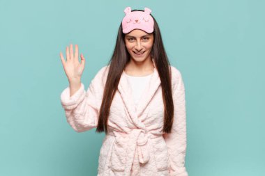 Genç ve güzel bir kadın mutlu ve neşeli bir şekilde gülümsüyor, el sallıyor, sizi selamlıyor ve selamlıyor, ya da veda ediyor. Pijama giyip uyanmak