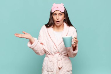 Genç ve güzel bir kadın şaşırmış ve şok olmuş görünüyor. Çenesi düşmüş ve eli açık bir objeyi tutuyor. Pijama giyip uyanmak