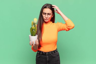 Genç, güzel bir kadın şaşkın ve kafası karışmış hissediyor. Kafasını kaşıyor ve yan tarafa bakıyor. Kaktüs kavramı