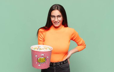 Genç ve güzel bir kadın mutlu bir şekilde gülümsüyor, eli kalçasında ve kendinden emin, olumlu, gururlu ve arkadaş canlısı bir tavırla. patlamış mısır konsepti