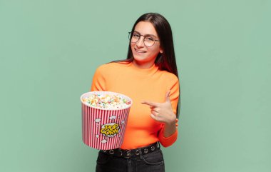 Genç, güzel bir kadın neşeyle gülümsüyor, mutlu hissediyor ve yan ve yukarı doğru işaret ediyor, fotokopi uzayında nesneler gösteriyor. patlamış mısır konsepti