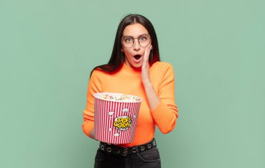 Genç ve güzel bir kadın şok olmuş ve korkmuş hissediyor, ağzı açık ve elleri yanaklarında korkmuş görünüyor. patlamış mısır konsepti