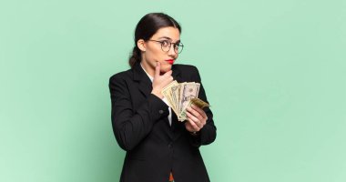 Genç, güzel bir kadın düşünüyor, kuşkulu ve şaşkın hissediyor, farklı seçeneklerle, hangi kararı vereceğini merak ediyor. iş ve banknotlar kavramı