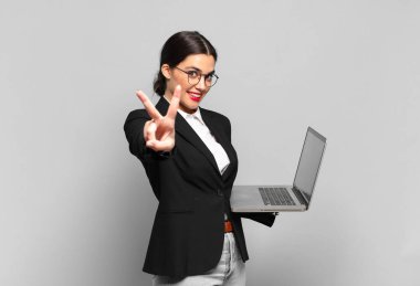 Genç ve güzel bir kadın gülümsüyor ve mutlu görünüyor, kaygısız ve pozitif, tek elle zafer ya da barış işareti yapıyor. dizüstü bilgisayar