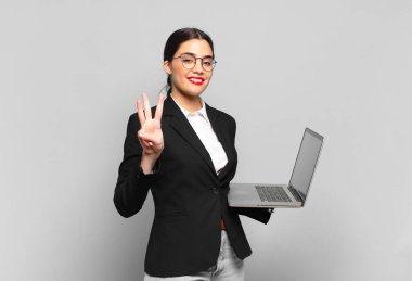 Genç, güzel bir bayan gülümsüyor ve arkadaş canlısı görünüyor. Üç ya da üç numarayı gösteriyor. Elleri önde, geriye doğru sayıyor. dizüstü bilgisayar