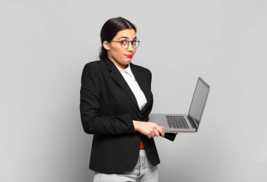 Genç, güzel bir kadın omuz silkiyor, kafası karışık ve kararsız, kolları bağlı ve şaşkın görünüyor. dizüstü bilgisayar