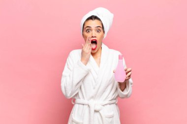 Genç ve güzel bir kadın şok olmuş ve korkmuş hissediyor, ağzı açık ve elleri yanaklarında korkmuş görünüyor. Bornoz ve yüz temizleme konsepti