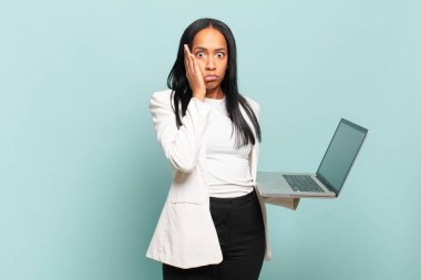 Genç siyahi kadın şok olmuş ve korkmuş hissediyor, ağzı açık ve elleri yanaklarında korkmuş görünüyor. dizüstü bilgisayar