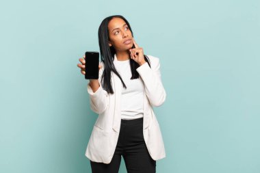 Genç siyahi kadın mutlu bir şekilde gülümsüyor ve hayal kuruyor ya da şüphe ediyor, yan tarafa bakıyor. akıllı telefon kavramı