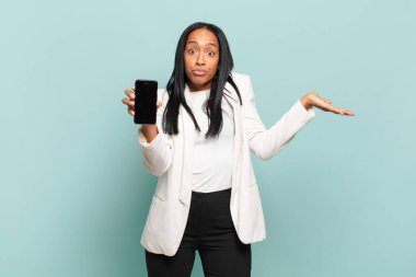 Genç siyahi kadın şaşkın ve şaşkın hissediyor, şüpheci, farklı seçenekleri tartıyor ya da komik bir ifadeyle seçiyor. akıllı telefon kavramı