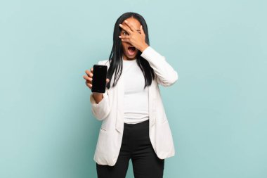 Genç siyahi kadın şok olmuş, korkmuş ya da korkmuş görünüyordu. Yüzü elle kaplıydı ve parmaklarının arasından bakıyordu. akıllı telefon kavramı