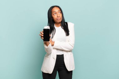 Genç siyahi kadın omuz silkiyor, kafası karışık ve kararsız, kollarını kavuşturup şaşkın bakışlarla kuşkulanıyor. akıllı telefon kavramı