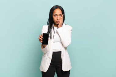 Genç siyahi kadın şok olmuş ve korkmuş hissediyor, ağzı açık ve elleri yanaklarında korkmuş görünüyor. akıllı telefon kavramı