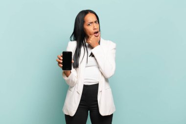 Ağzı ve gözleri açık, eli çenesinde genç siyah bir kadın. Hoş olmayan bir şok geçiriyor. Ne diyor? akıllı telefon kavramı